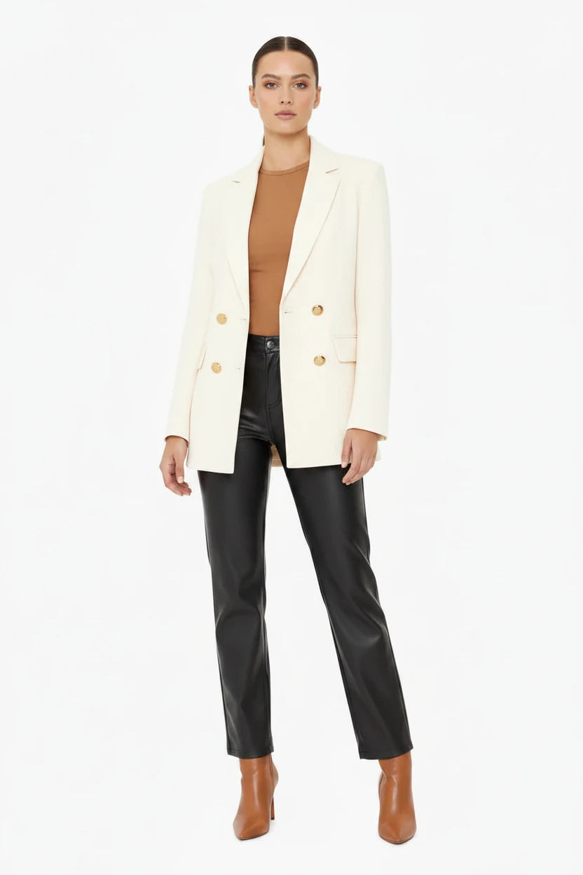 A model stands against a plain white studio background wearing a cream-colored blazer over a brown ribbed top, black leather-look pants, and brown ankle boots.